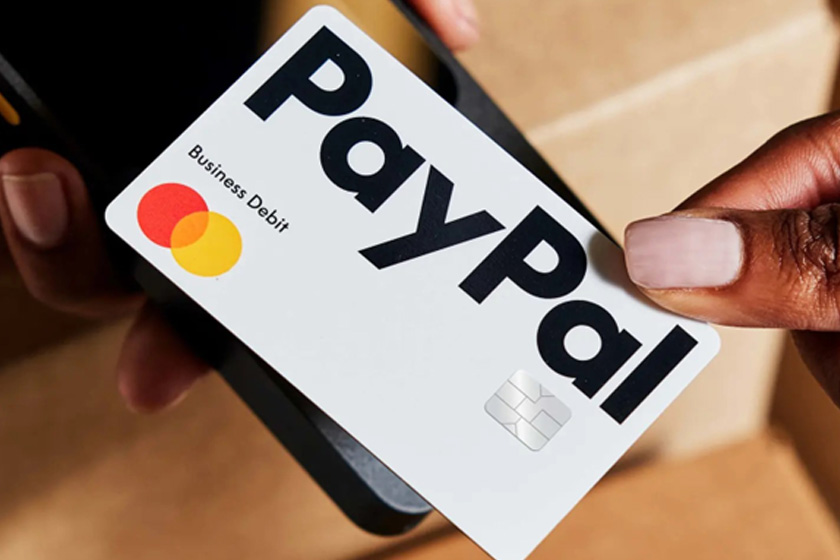Eine Person hält eine PayPal Business Debit Mastercard auf ein Kartenlesegerät oder Smartphone. Copyright : PayPal
