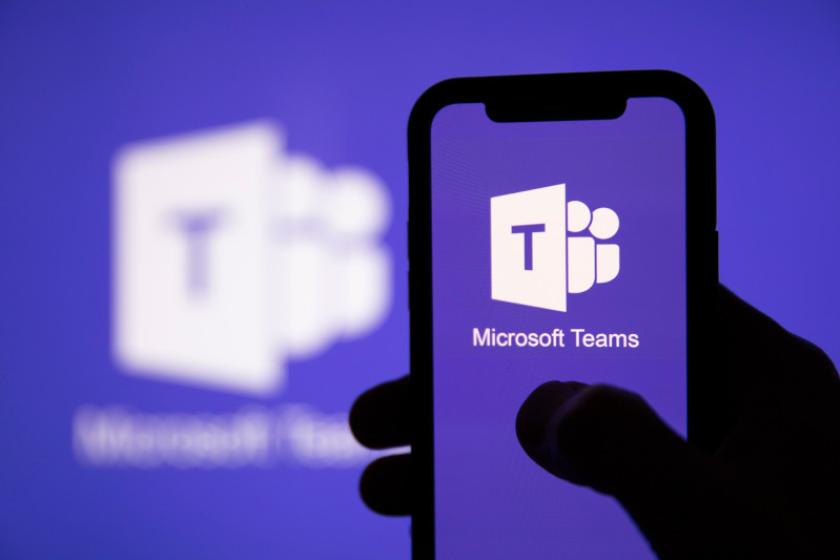 Ein Smartphone mit Microsoft-Teams-Logo auf dem Display wird in einer Hand gehalten, dahiner leuchtet ebenfalls ein Teams-Logo.