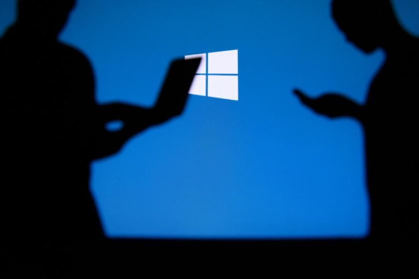 Schatten von Menschen, die mobile Devices bedienen, vor einem großen Windows-Logo auf blauem Hintergrund.