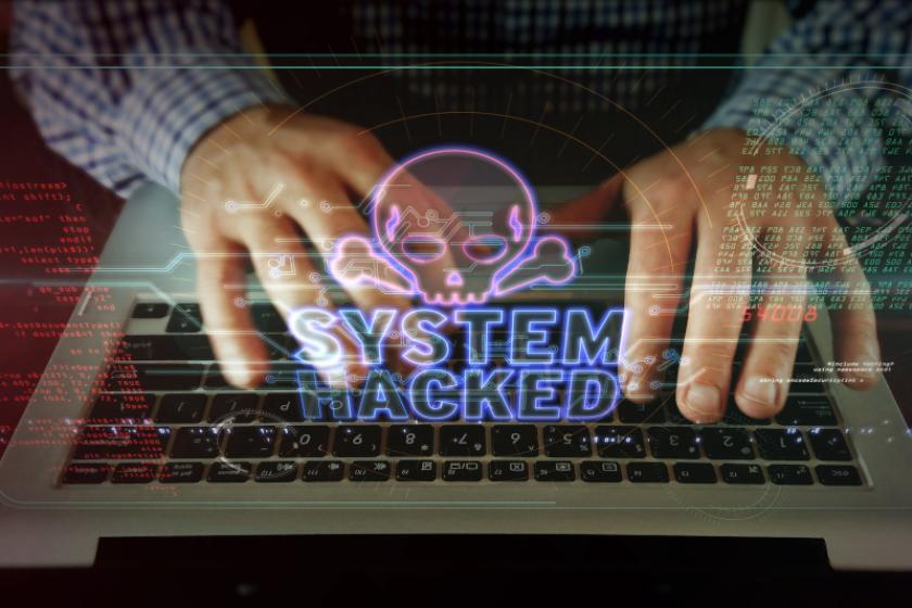 Zwei Hände, die auf einer Laptop-Tastatur tippen. Darüber schwebt ein Totenkopf mit dem Schriftzug "System Hacked".