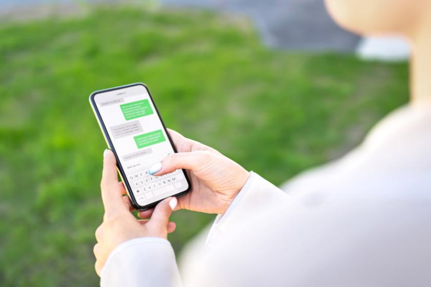 Frau blickt draußen auf einer Wiese auf einen Chatverlauf auf ihrem Smartphone, über die Schulter fotografiert..