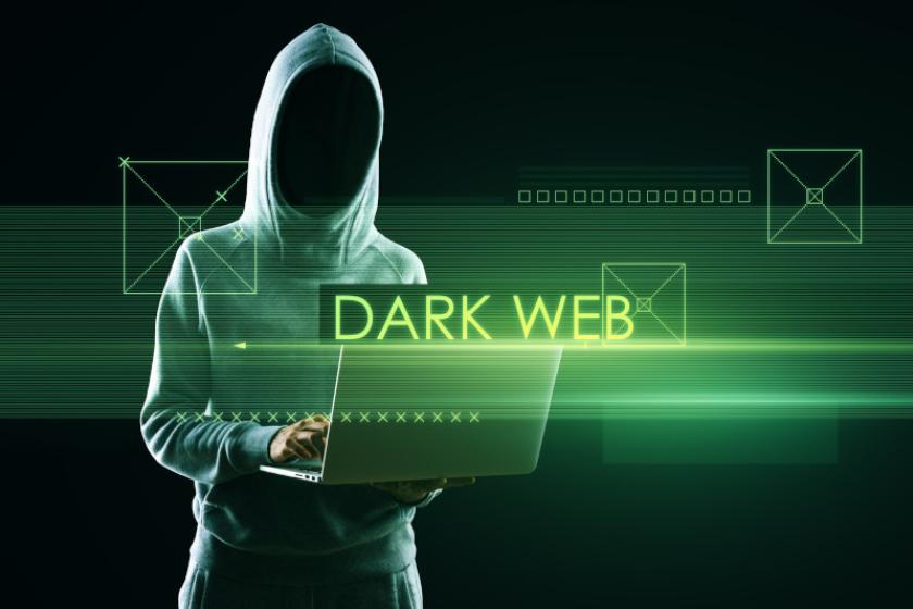 Grün angeleuchtete Person in grauem Kapuzenpulli und nicht erkennbarem Gesicht steht mit einem Laptop in der Hand, davor steht "Dark Web"