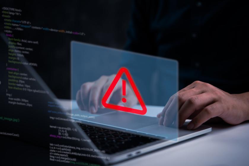 Ein Warnschild mit rotem Ausrufezeichen darin schwebt über einem geöffneten Laptop, dahinter befindet sich Programmiercode.