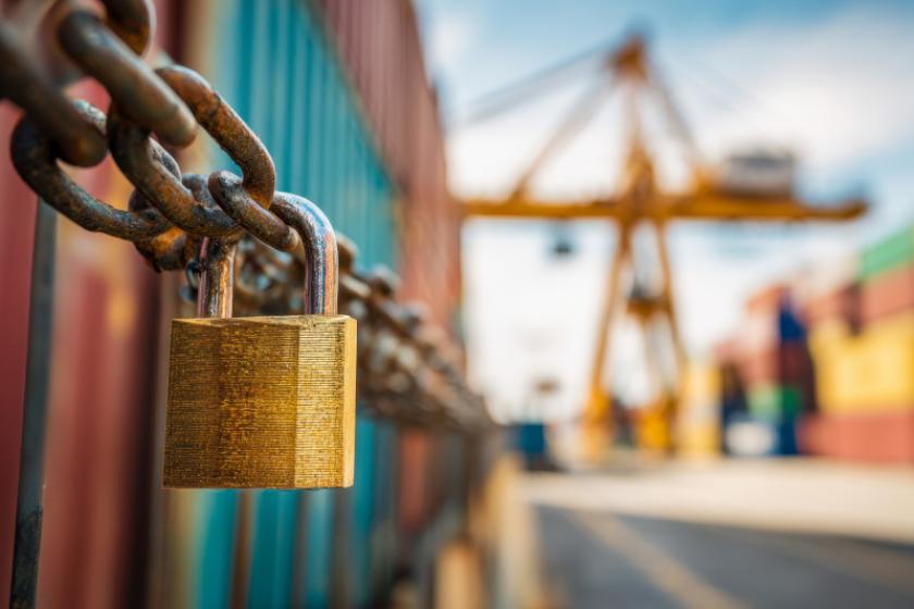 Eine Kette mit Vorhängeschloss umschließt mehrere Schiffscontainer, im Hintergrund eine Container-Hafen-Szenerie mit gelbem Kran.
