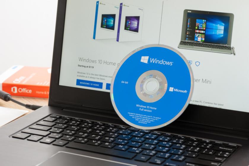 Eine blaue Installations-CD für Windows 10 lehnt an einem geöffneten und eingeschalteten Laptop, auf dem Informationen zu Windows 10 angezeigt werden.