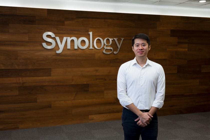 Ein Herr in weißem Hemd steht vor einer Holzwand mit dem Synology Logo darauf