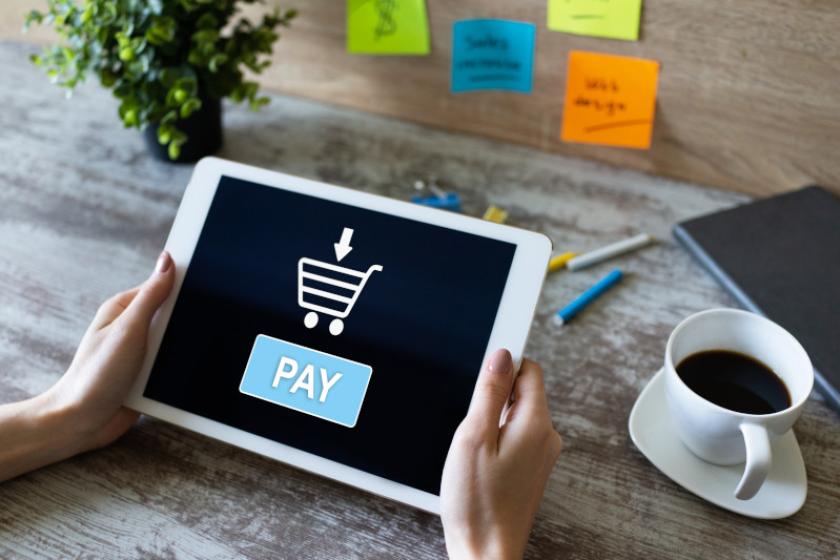 Person hält ein Tablet über einem Tisch neben einer Kaffeetasse, auf dem Display steht "Pay" sowie ein Einkaufswagensymbol.