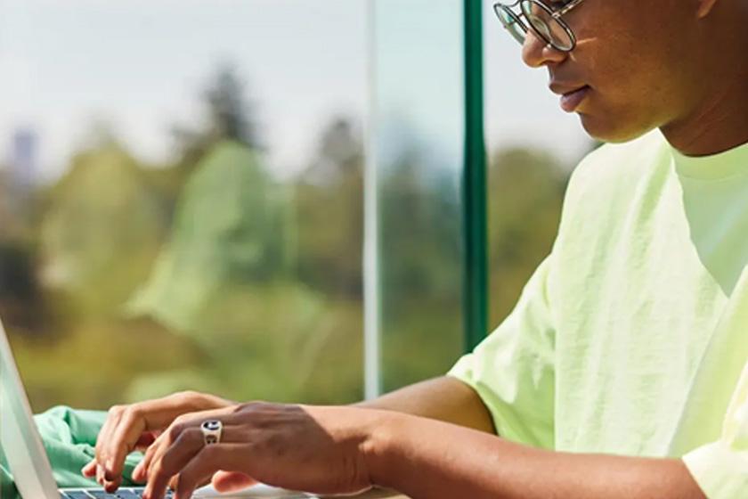 Person tippt in Laptop, im Hintergrund schaut man durch Fenster ins Gründe