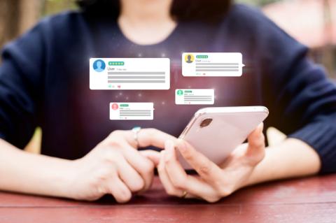 Frau sitzt an einem Tisch und blickt auf ihr Smartphone, über dem Chatblasen schweben.