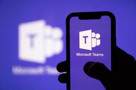 Ein Smartphone mit Microsoft-Teams-Logo auf dem Display wird in einer Hand gehalten, dahiner leuchtet ebenfalls ein Teams-Logo.