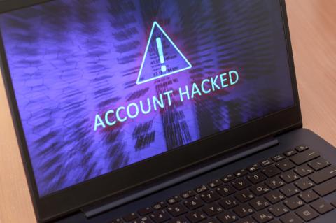 Schwarzer aufgeklappter Laptop mit einem violett hinterlegten Ausrufezeichen und dem Schriftzug Account Hacked auf dem Bildschirm.