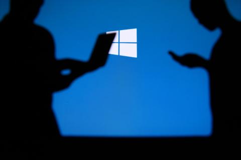 Schatten von Menschen, die mobile Devices bedienen, vor einem großen Windows-Logo auf blauem Hintergrund.
