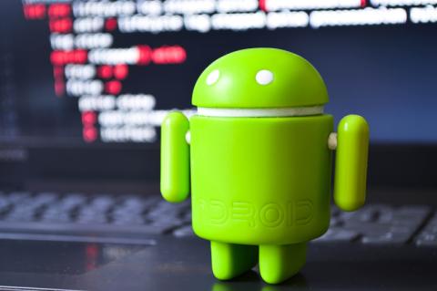 Ein grüner Android-Robotoer steht auf einem aufgeklappten Laptop, auf dessen Bildschirm Prorgammiercode steht.