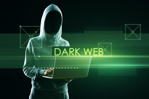 Grün angeleuchtete Person in grauem Kapuzenpulli und nicht erkennbarem Gesicht steht mit einem Laptop in der Hand, davor steht "Dark Web"