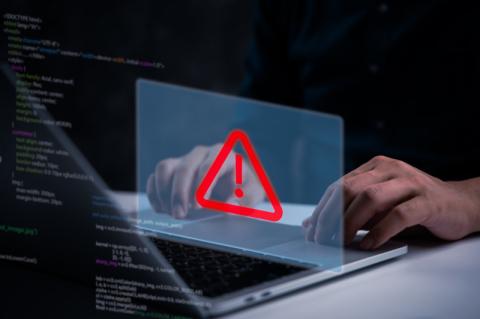 Ein Warnschild mit rotem Ausrufezeichen darin schwebt über einem geöffneten Laptop, dahinter befindet sich Programmiercode.
