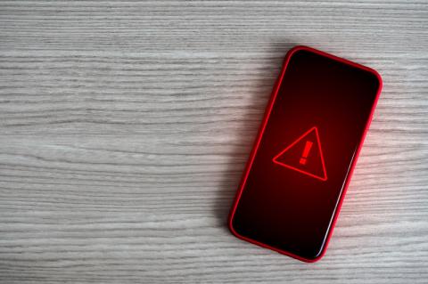Ein Smartphone liegt auf einer Holzoberfläche und zeigt ein rotes Ausrufezeichen in einem roten Dreieck auf schwarzem Hintergrund an.