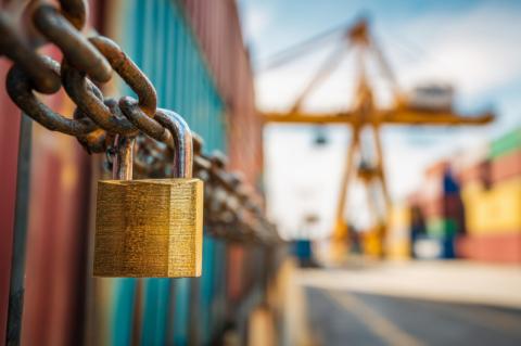 Eine Kette mit Vorhängeschloss umschließt mehrere Schiffscontainer, im Hintergrund eine Container-Hafen-Szenerie mit gelbem Kran.