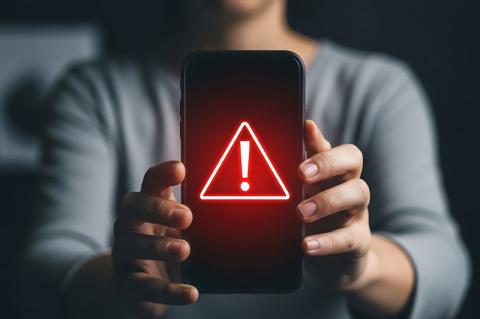 Ein Mann hält ein Smartphone mit beiden Händen hoch, auf dem ein rotes Warndreieck mit Ausrufezeichen darin auf dem Display erscheint.