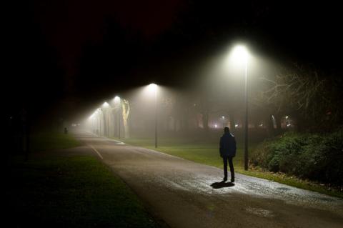 Eine dunkel gekleidete Person geht bei Nacht auf einem von Straßenlampen beleuchteten Weg spazieren.