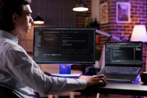 Ein Mann sitzt an einem Laptop sowie einem weiteren Bildschirm daneben, auf denen Programmiercode abgebildet ist.