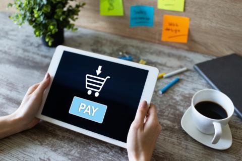 Person hält ein Tablet über einem Tisch neben einer Kaffeetasse, auf dem Display steht "Pay" sowie ein Einkaufswagensymbol.