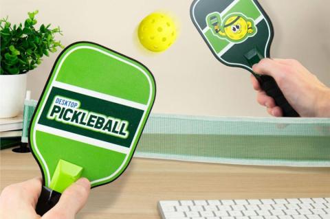 Zwei Hände spielen Pickleball auf einem Schreibtisch mit Mini-Netz und kleinen Holzschlägern – das kompakte Set sorgt im Büro für Bewegung und Spaß.(Quelle: monsterzeug.de)