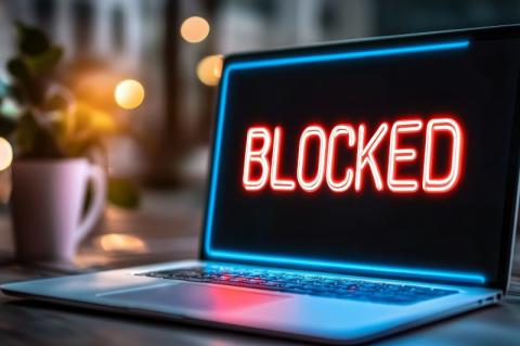 Ein aufgeklappter Laptop steht auf einem Tisch, auf dem ansonsten schwarzen Bildschirm steht in roten Buchstaben "BLOCKED".