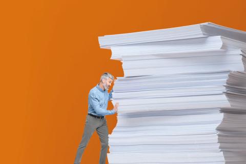Ein Mann mit grauen Haaren und blauem Hemd steht vor einem überdimensionalen Papierstapel auf orangem Hintergrund, der sinnbildlich für Bürokratie und Überlastung wirkt.