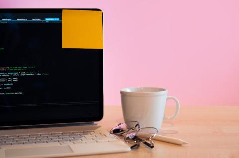Ein aufgeklappter Laptop mit Programmiercode auf dem Bildschirm steht auf einem Tisch. Daneben steht eine Kaffeetasse und liegt ein Schlüsselbund. Am Bildschirm klebt oben rechts ein Post-it.