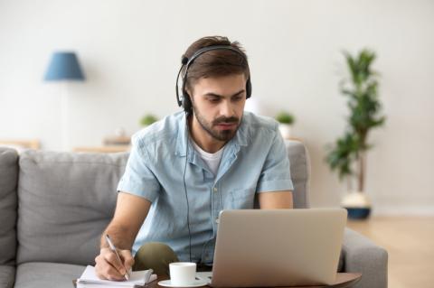 Ein junger Mann sitzt mit Headset vor einem geöffneten Notebook und schaut auf den Bildschirm.